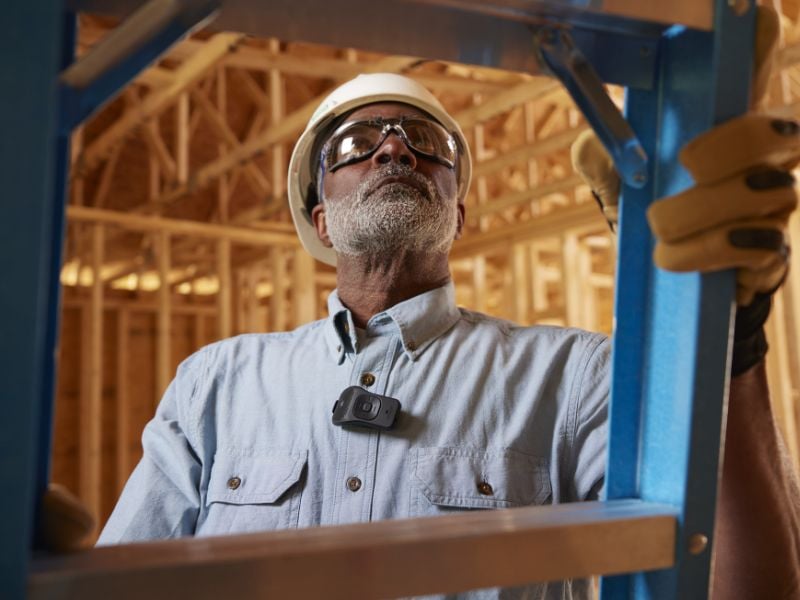 A man climbs a ladder wearing a hard hat. The man has a Noxgear 39g speaker clipped to his button-down work shirt showing how easily the speaker can be attached for use at the job site.