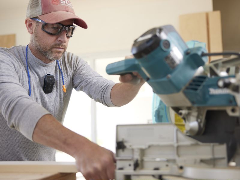 A man working with a miter saw wears safety glasses and a ballcap. He has a Noxgear 39g attached to his shirt using the Magnetic clip.
