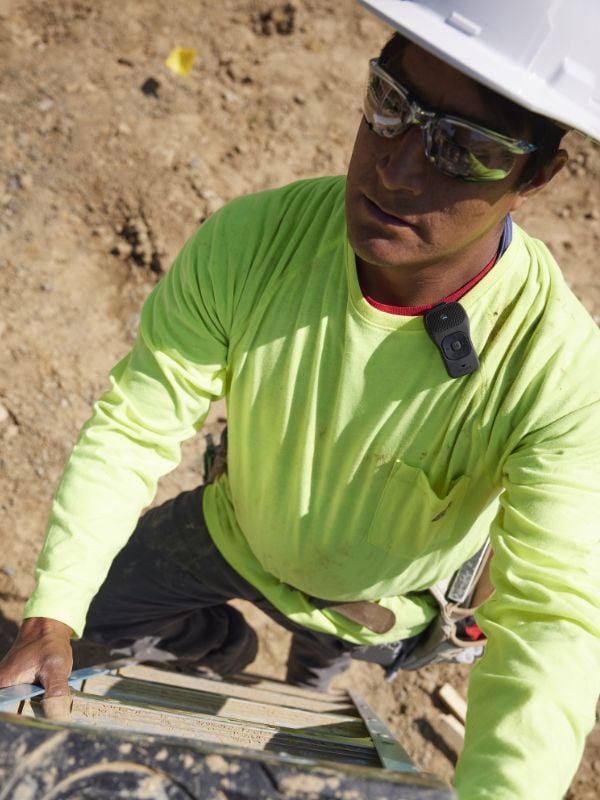A man is climbing a ladder while wearing a hard hat and safety glasses. His Noxgear 39g speaker is attched to the neckline of his shirt with the Sport clip. This makes it easy to attach to many clothing items while working.