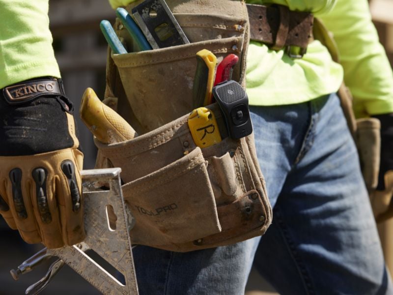 A Noxgear 39g speaker is clipped onto a leather toolbelt on a person's hip. The speaker is ideal for job sites.