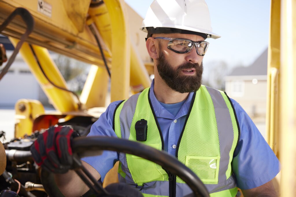 A man is operating heavy machinery at a jobsite while wearing the Noxgear 39g speaker attached to his safety vest using the Magnetic clip on the speaker. This clip makes the speaker ideal for construction workers and others working at job sites.
